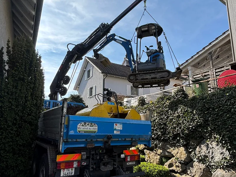 Kranwagen hebt einen Minibagger über die Gartenmauer